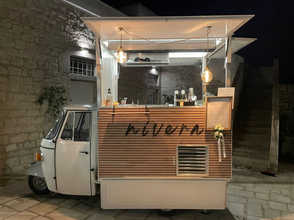 Ape Car Nìvera vista frontalmente con gli sportelli aperti, bancone attrezzato con bicchieri e bottiglie, drink list e rose bianche, davanti a un muro in pietra