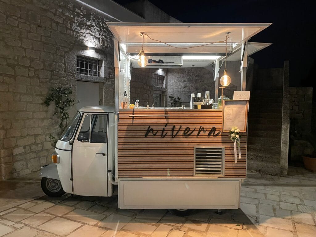 Ape Car Nìvera vista frontalmente di sera, bancone in legno a doghe con la scritta nivera, lampadine a filamento accese, drink list incorniciata e rose bianche