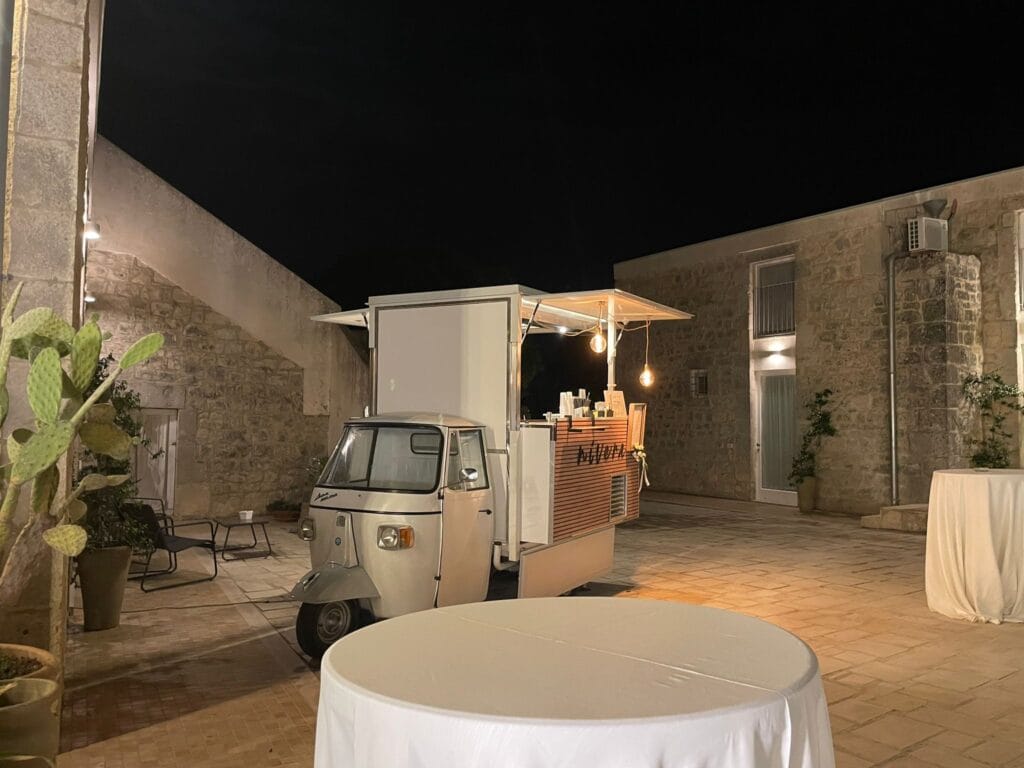 Ape Car Nìvera illuminata di sera in un cortile in pietra siciliano, con fichi d'India in vaso e tavoli alti vestiti di bianco