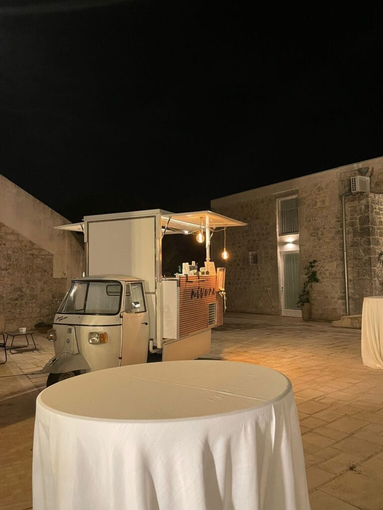 Ape Car Nìvera illuminata in fondo a un cortile in pietra siciliano, con un tavolo alto vestito di bianco in primo piano