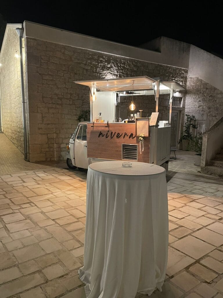Ape Car Nìvera illuminata accanto a un lungo muro in pietra, con un tavolo alto vestito di bianco in primo piano su pavimentazione in pietra