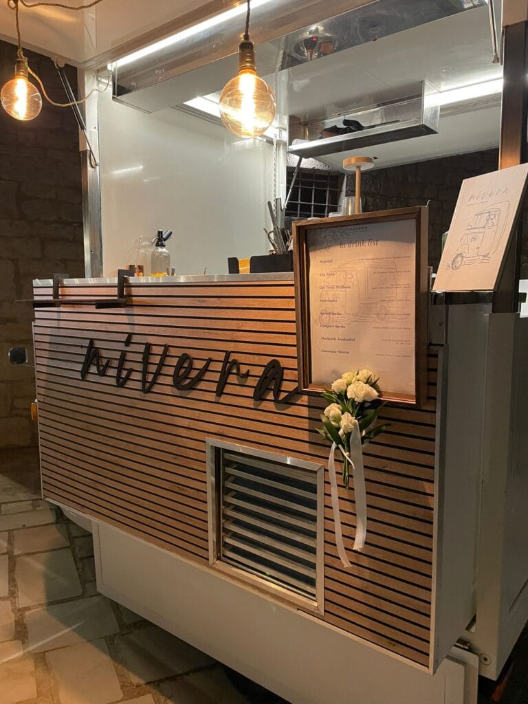 Dettaglio ravvicinato del bancone in legno a doghe dell'Ape Nìvera con la scritta in corsivo, la drink list incorniciata, rose bianche e lampadine a filamento