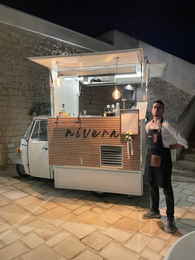 Bartender Nìvera in grembiule accanto all'Ape Car illuminata di sera, con drink list incorniciata e rose bianche sul bancone