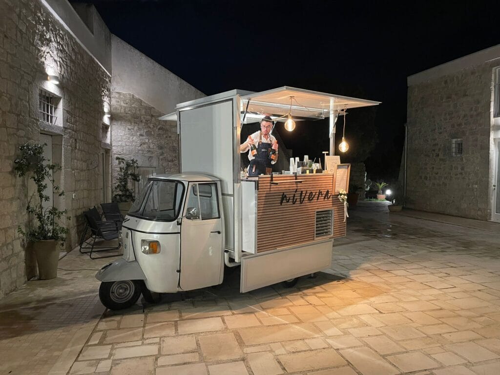 Bartender Nìvera che impugna due bottiglie di Aperol al bancone dell'Ape Car illuminata, in un cortile in pietra di sera