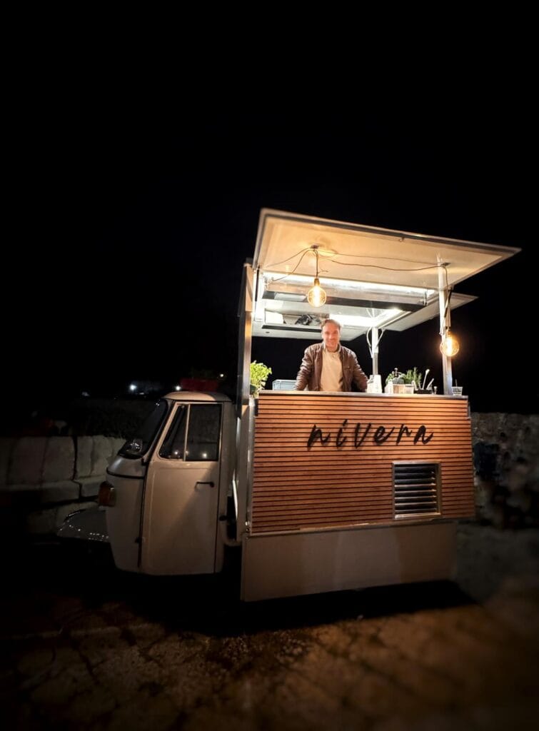 Bartender Nìvera dietro il bancone in legno dell'Ape Car illuminato da lampadine a filamento, di sera, davanti a un muro in pietra