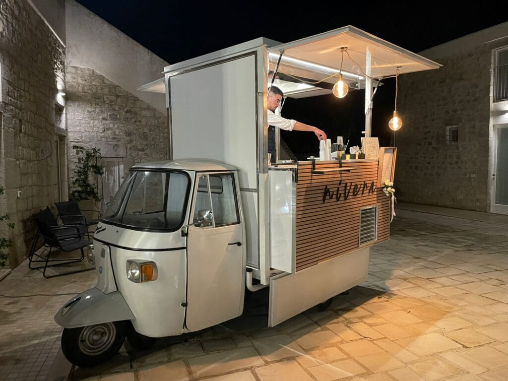 Bartender Nìvera che prepara il servizio al bancone dell'Ape Car in un cortile in pietra illuminato di sera
