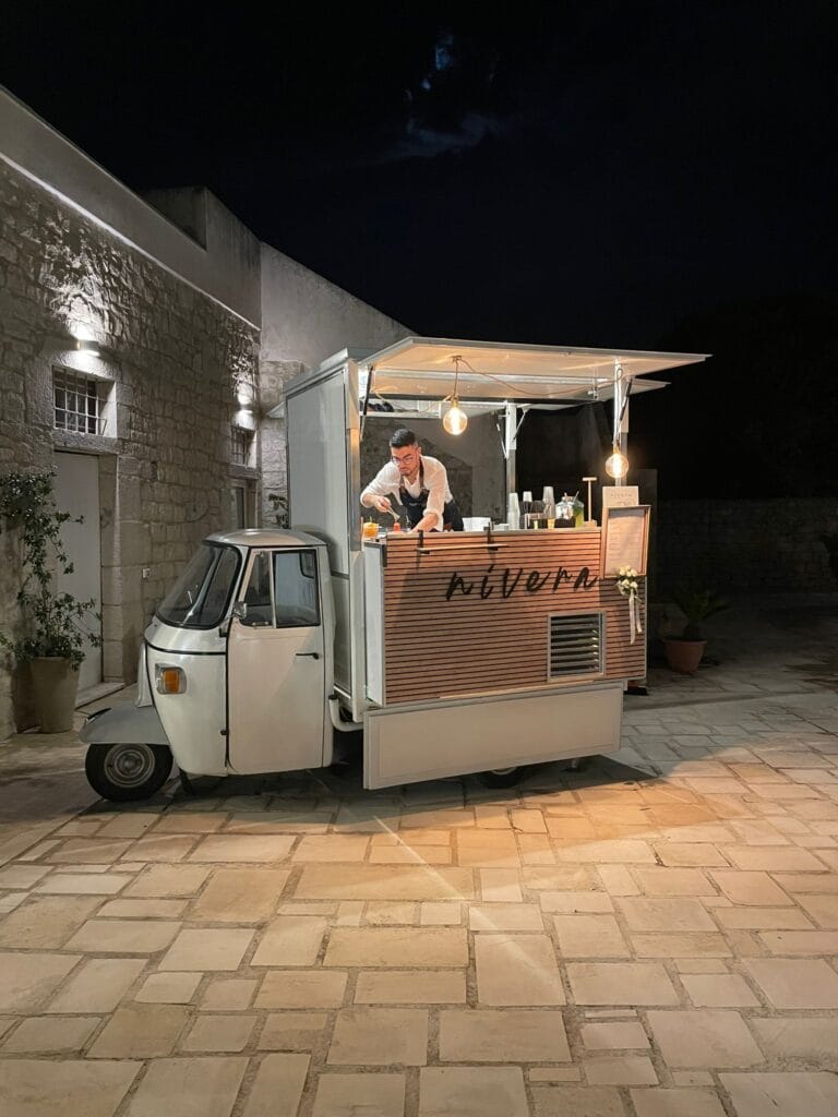 Bartender Nìvera che prepara i cocktail al bancone dell'Ape Car illuminata di sera in un cortile in pietra