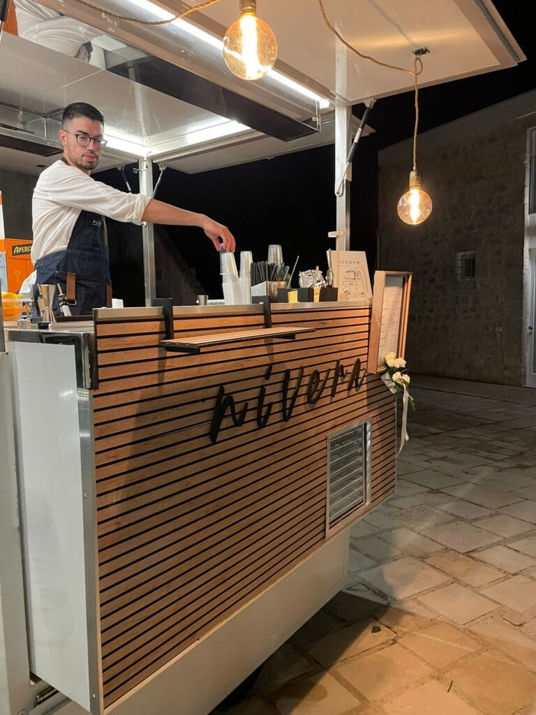 Bartender Nìvera che prende dei bicchieri dal bancone dell'Ape Car, con lampadine a filamento e drink list incorniciata