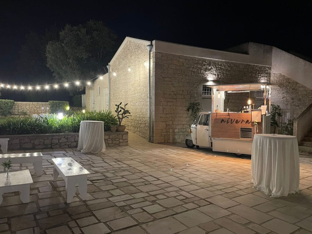 Ampio cortile in pietra di una masseria siciliana allestito con luci a festone, panche bianche e l'Ape Car Nìvera illuminata sul fondo