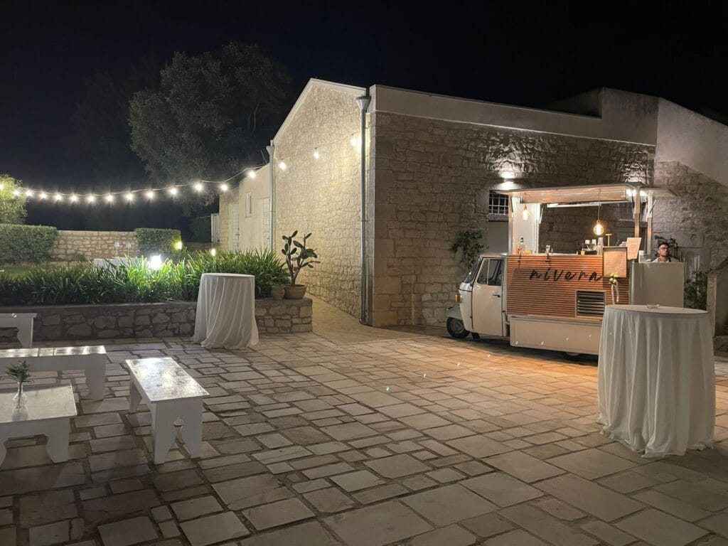Cortile di masseria allestito per un ricevimento con luci a festone, panche bianche, tavoli alti e l'Ape Car Nìvera illuminata sullo sfondo