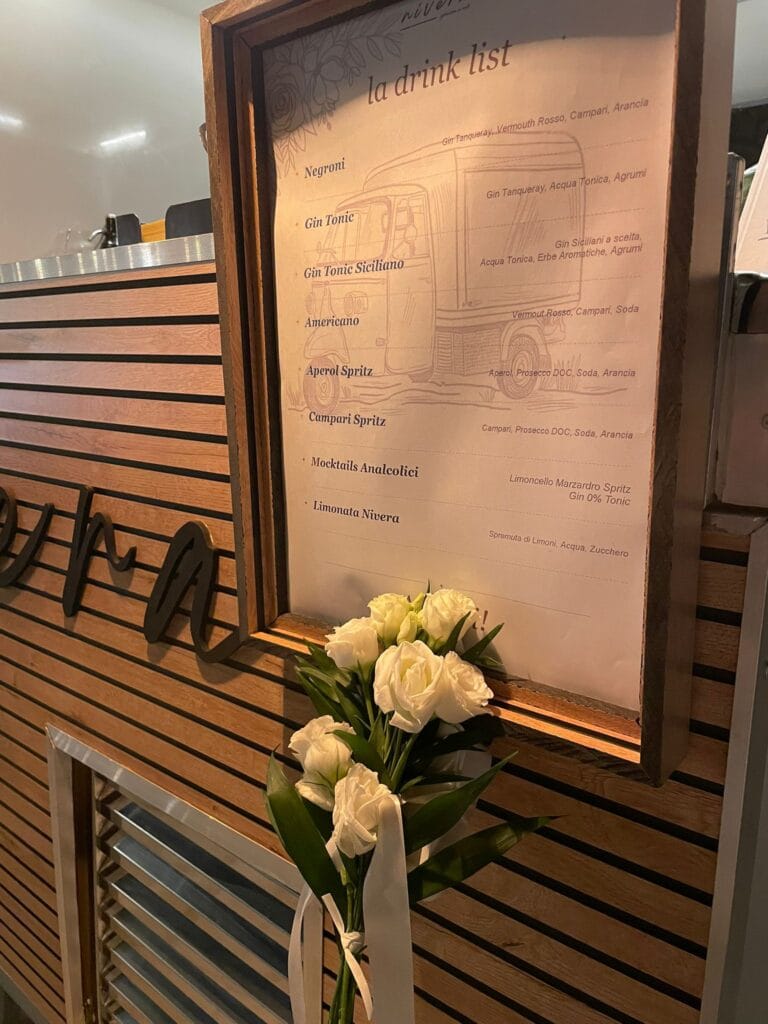 Dettaglio della drink list Nìvera incorniciata con Negroni, Gin Tonic Siciliano, Aperol Spritz, mocktail analcolici e limonata, accanto a un mazzo di rose bianche