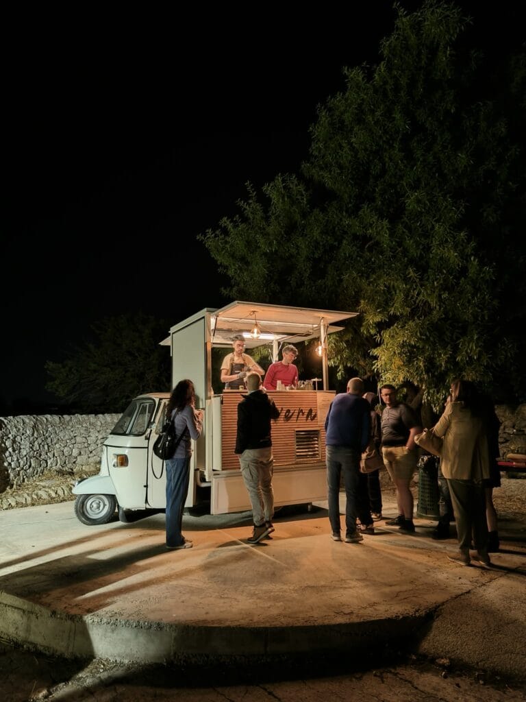 Gruppo di ospiti servito al bancone dell'Ape Car Nìvera durante un aperitivo serale all'aperto, con muretto a secco e alberi sullo sfondo