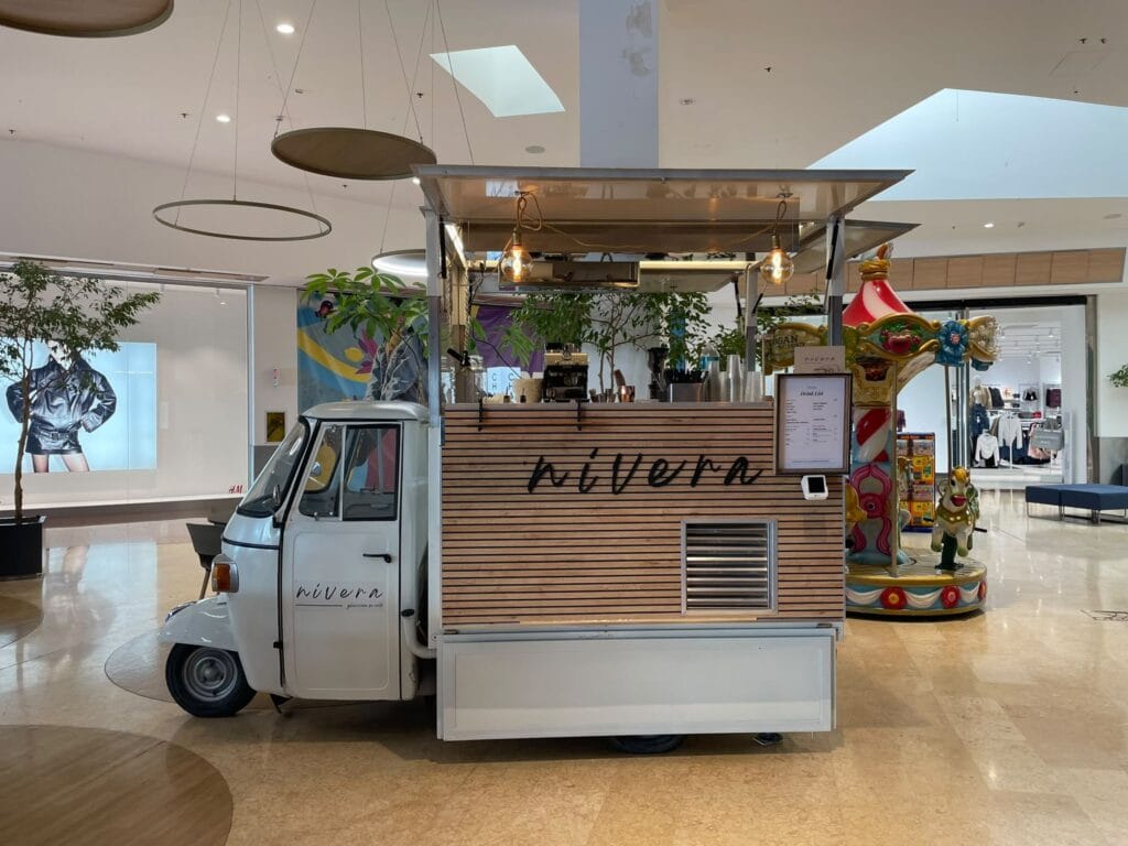 Ape Car Nìvera vista frontalmente nella galleria di un centro commerciale, con bancone in legno attrezzato con macchina del caffè, bicchieri e drink list incorniciata